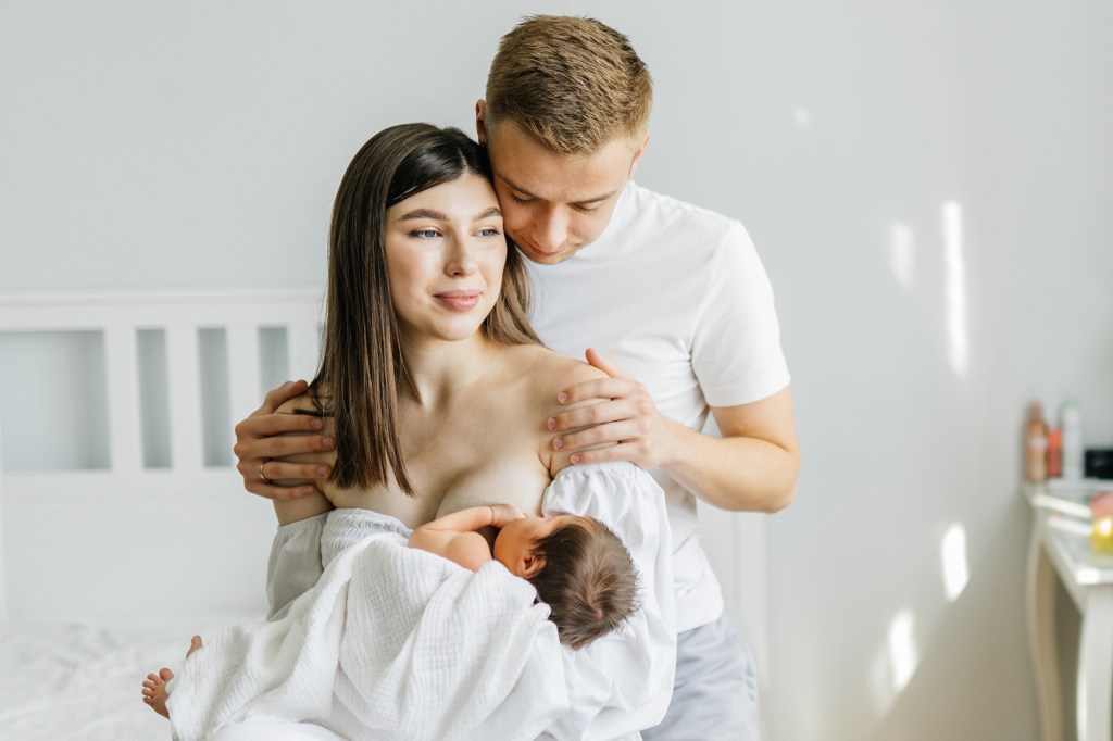 Couple in love with similar Mario Novembre, sitting in a cozy bedroom; woman breastfeeding her newborn daughter while her husband gives her a warm hug.